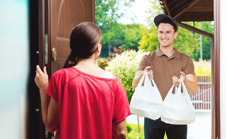 Young delivery man delivering Chinese take away food for young woman, standing at the entrance door and holding two plastic bags in hands.