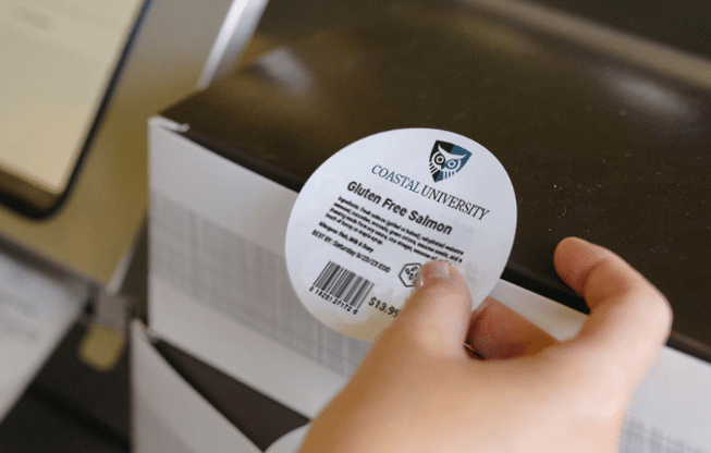 A food service worker applying a custom circular nutrition label to a meal container, demonstrating automated labeling for school cafeteria compliance